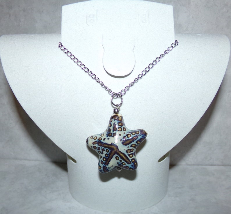 Puede incluir: Un collar de cadena plateada con un colgante de estrella de mar de cer&aacute;mica blanca. La estrella de mar tiene un esmalte marr&oacute;n y azul con un patr&oacute;n moteado.