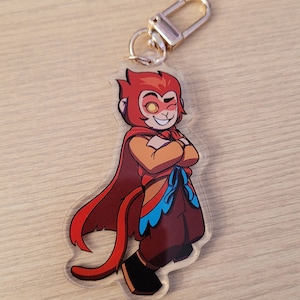 May include: A clear acrylic keychain featuring a cartoon character with red hair, a red cape, and brown pants. The character has a confident expression and is wearing a gold-colored clasp.