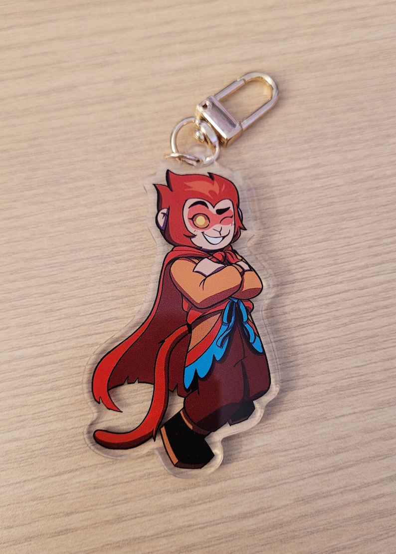 May include: A clear acrylic keychain featuring a cartoon character with red hair, a red cape, and brown pants. The character has a confident expression and is wearing a gold-colored clasp.