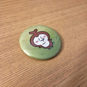 Könnte beinhalten: Ein runder Button mit einem Cartoon-Charakter-Design. Der Button zeigt einen braunen Charakter mit weißem Gesicht und rosa Wangen auf grünem Hintergrund. Der Button hat einen Durchmesser von etwa 5 cm.
