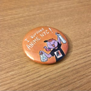 May include: Orange button with the text "I survived Anime NYC!" and a cartoon character holding a drink and a bag of snacks. The character wears a black shirt with a logo and a red cap.