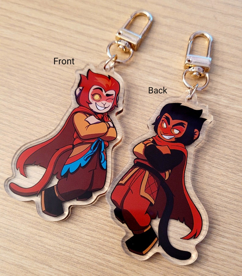 May include: Clear acrylic keychain featuring a cartoon monkey character with a red and orange color scheme. The front and back of the charm are visible, each with a different design. The keychain has a gold-colored clasp.