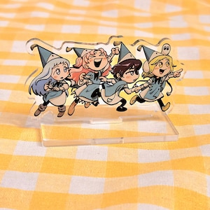 May include: A clear acrylic standee featuring a colorful illustration of four cartoon characters wearing blue pointed hats and matching outfits. The characters are depicted in a dynamic, running pose, with one character pointing forward.