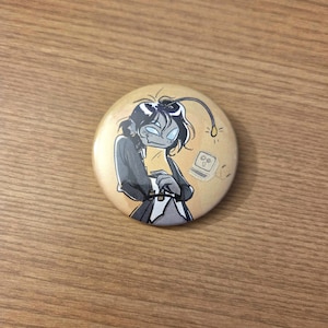 May include: A round button with a cartoon illustration of a person with a lightbulb antenna and a laptop. The person is wearing a dark gray jacket and a white shirt. The background is a light tan color.