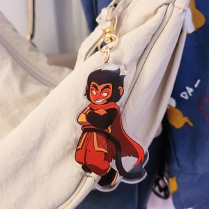 May include: A clear acrylic keychain featuring a cartoon character with a mischievous grin. The character has black hair, a red cape, and is wearing red and gold clothing. The keychain is attached to a gold-colored clasp.