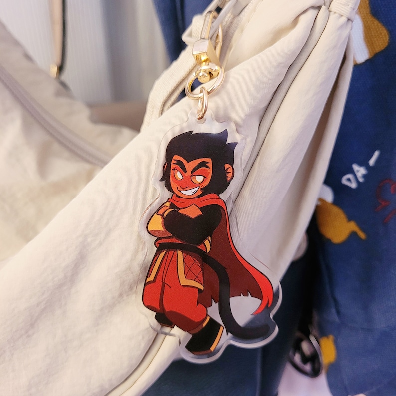 May include: A clear acrylic keychain featuring a cartoon character with a mischievous grin. The character has black hair, a red cape, and is wearing red and gold clothing. The keychain is attached to a gold-colored clasp.