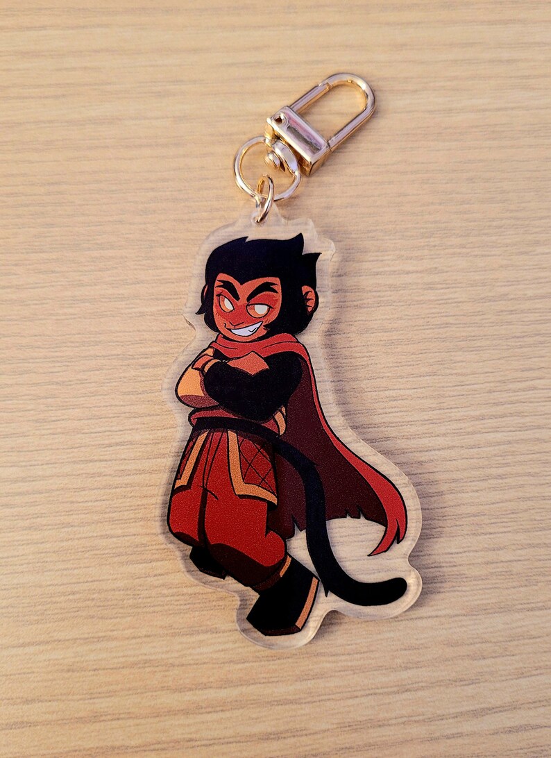 May include: A clear acrylic keychain featuring a cartoon character with a red and black color scheme. The character has crossed arms and a confident expression. The keychain has a gold-colored clasp.
