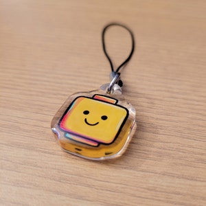 May include: A clear acrylic keyring with a yellow suitcase design and a smiling face. The suitcase has a colourful outline and a black string for attachment. The keyring is on a wooden surface.