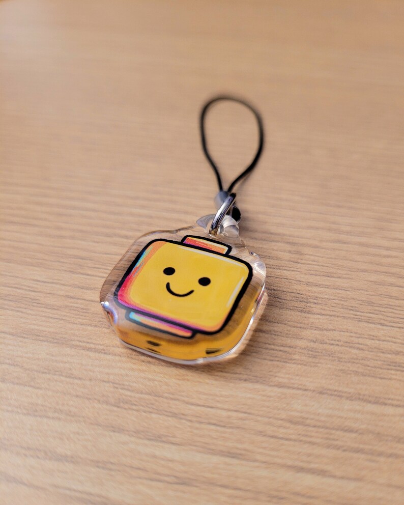 May include: A clear acrylic keyring with a yellow suitcase design and a smiling face. The suitcase has a colourful outline and a black string for attachment. The keyring is on a wooden surface.