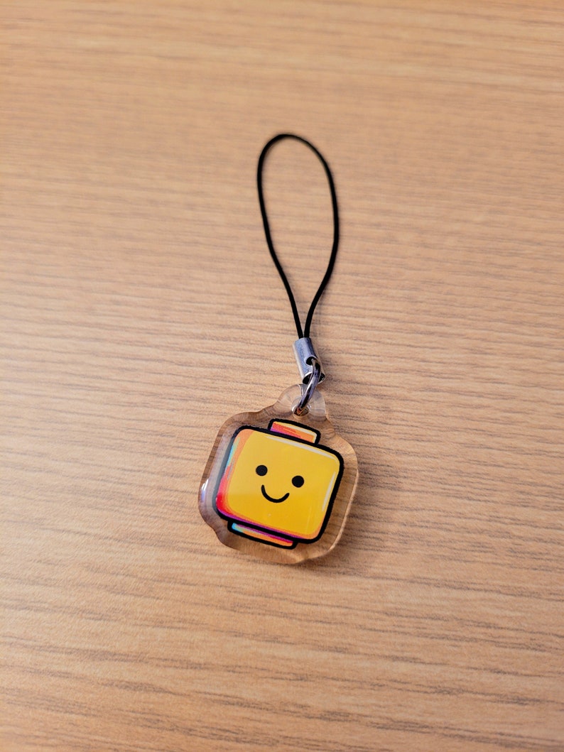 May include: A clear acrylic charm depicting a yellow Lego-style head with a smiling face and a 3D effect in red, pink and blue. The charm is attached to a black cord with a silver clasp, suitable for use as a keyring or phone charm.