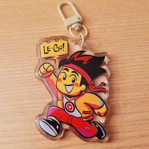 May include: A clear acrylic keychain featuring a cartoon character in a running pose. The character wears a red headband, red pants, and a yellow jacket. The keychain has a metal clasp and the text "LE-GO!" in a yellow speech bubble.