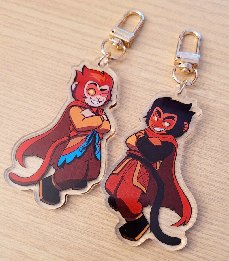 May include: Two acrylic keychains featuring cartoon characters with red and orange color schemes. Each keychain has a gold-colored clasp. The characters are depicted with crossed arms and capes, with one having a tail. The background is a light wood surface.