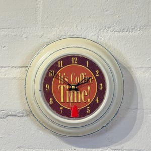 May include: A round wall clock with a distressed cream-coloured frame. The clock face features a dark red centre with the text 'It's Coffee Time!' in gold lettering, a small red coffee pot illustration, and black clock hands with gold numerals.