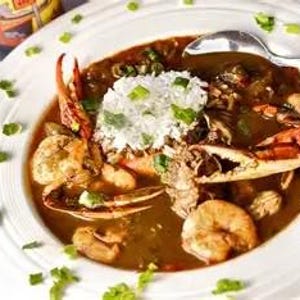 May include: A white bowl filled with seafood gumbo, featuring crab claws, shrimp, and rice. The stew is a deep brown color, garnished with green herbs, and served with a spoon.