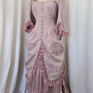 Op de afbeelding: Een oudroze jurk met een vierkante halslijn, een knoopsluiting aan de voorzijde en driekwartmouwen met kanten ruches. Het kledingstuk is voorzien van een bloemrijk jacquardpatroon, een stoffen bloemaccent en een gelaagde rok met kanten afwerking.