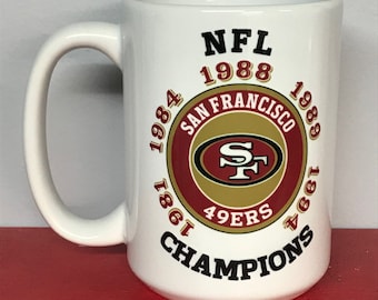 Op de afbeelding: Een witte keramische mok met een rood, goud en zwart San Francisco 49ers logo. De mok heeft de tekst "NFL 1988 1984 SAN FRANCISCO 1989 1986 49ERS CHAMPIONS" rond het logo.