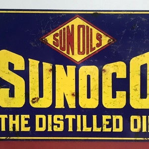 May include: Vintage metal sign advertising Sunoco petrol. The sign is blue with yellow lettering and a red and yellow diamond logo that says "Sun Oils". The text on the sign reads "Sunoco The Distilled Oil".