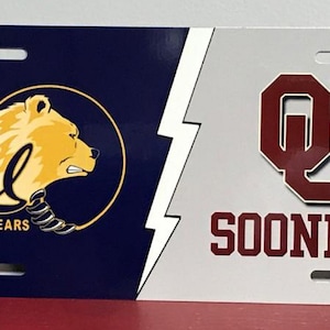 May include: A white and blue license plate with a gold bear and the text "Cal Golden Bears" on the left side. The right side of the plate is white with a maroon "OU" logo and the text "Sooners".