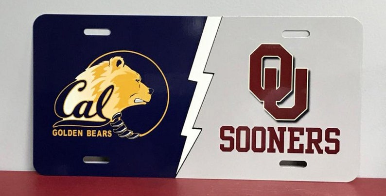 May include: A white and blue license plate with a gold bear and the text "Cal Golden Bears" on the left side. The right side of the plate is white with a maroon "OU" logo and the text "Sooners".