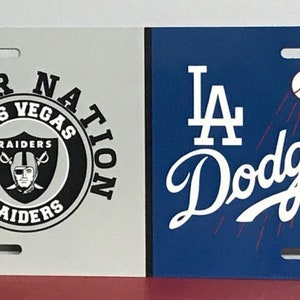 May include: A white and blue license plate with two team logos. The left side features a black and white logo for the Las Vegas Raiders with the text "Raider Nation Las Vegas Raiders". The right side features a blue and white logo for the Los Angeles Dodgers with the text "LA Dodgers".