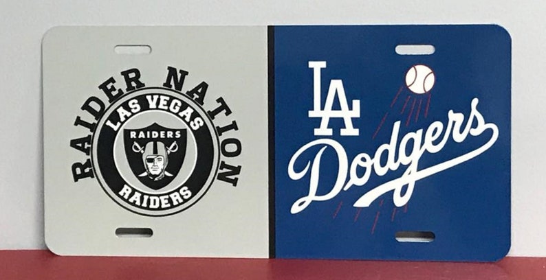 May include: A white and blue license plate with two team logos. The left side features a black and white logo for the Las Vegas Raiders with the text "Raider Nation Las Vegas Raiders". The right side features a blue and white logo for the Los Angeles Dodgers with the text "LA Dodgers".