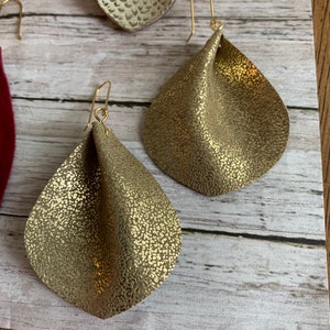 May include: A pair of gold-toned dangle earrings with a textured, crinkled leather design. The earrings have a simple, minimalist style.