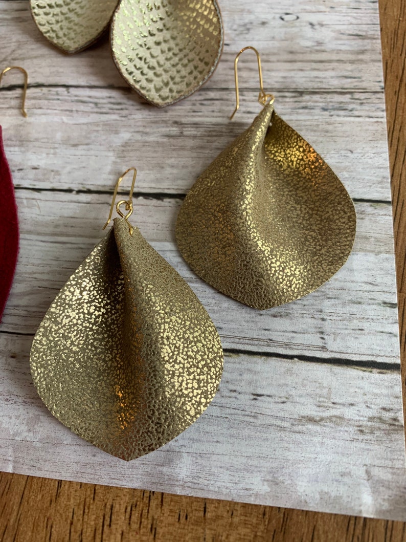 May include: A pair of gold-toned dangle earrings with a textured, crinkled leather design. The earrings have a simple, minimalist style.