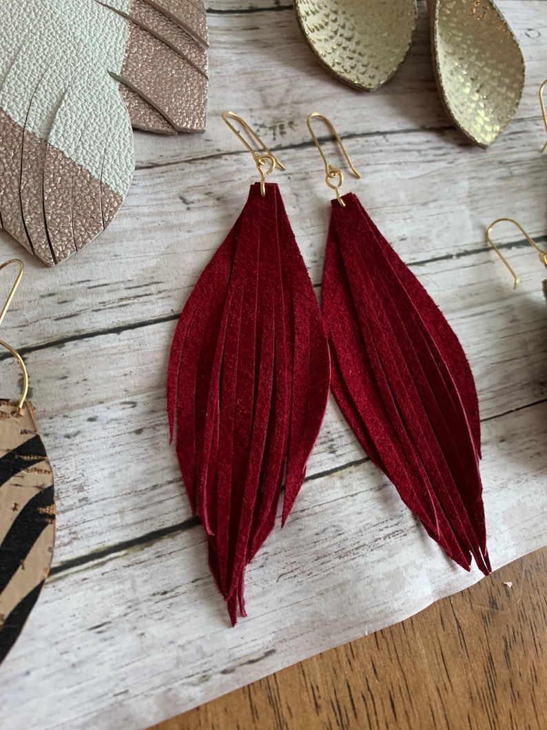May include: A pair of burgundy leather tassel earrings with gold hooks. The earrings are long and have a fringe design.