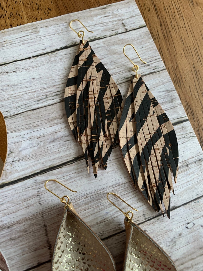 May include: A pair of long, dangling earrings with a black and tan zebra print pattern. The earrings are made of leather and have a gold-tone hook closure.