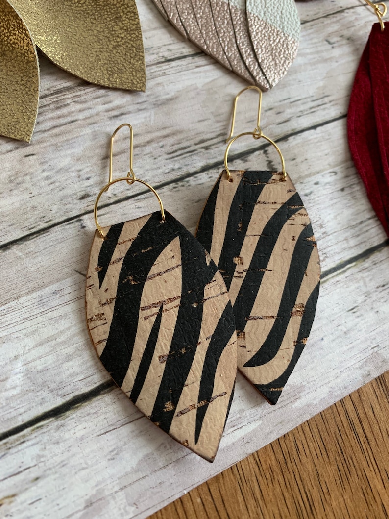 May include: A pair of cork earrings with a black and white zebra print. The earrings are shaped like a teardrop and have gold hooks.
