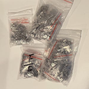 May include: Four clear plastic bags filled with small, silver-colored metal charms. The charms appear to be letters and numbers. The bags are sealed with red zippers and are arranged on a white surface.