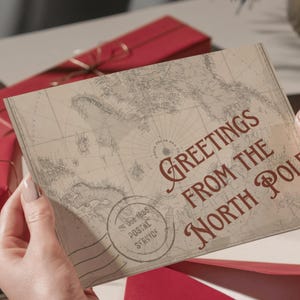 May include: A vintage-style greeting card with the words "Greetings From The North Pole" in burgundy script. The card features a map design and a postal service stamp. The card is held by a person.