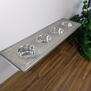 May include: A wooden pet feeder with four stainless steel bowls. The feeder is rectangular, stained gray, and mounted on a wall. The bowls are evenly spaced and appear to be for food or water. The background includes a wooden floor and greenery.
