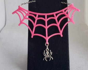 May include: Pink glitter spiderweb necklace with a silver spider charm. The necklace is on a black velvet display stand.