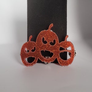 May include: Three orange glitter pumpkins with scary faces. The pumpkins are connected together and have a black background.