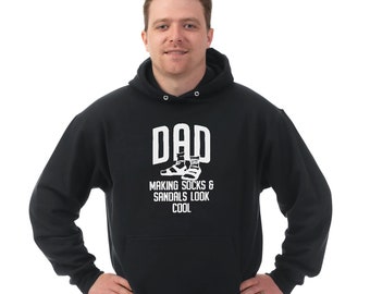 May include: Black hooded sweatshirt with white text that reads "DAD MAKING SOCKS & SANDALS LOOK COOL" and a graphic of a pair of sandals with socks on them.