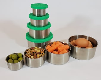 May include: A set of four stainless steel food storage containers with bright green silicone lids stacked vertically, alongside four open containers filled with green olives, mixed nuts, baby carrots, and brown eggs.