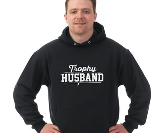 May include: Black hooded sweatshirt with the text "Trophy Husband" in white.