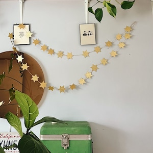 Könnte beinhalten: Zwei Girlanden aus goldenen Glitzersternen hängen an einer weißen Wand. Die Girlande ist zwischen zwei weißen Halterungen gespannt. Eine grüne Metall-Aufbewahrungsbox steht auf einer Holzoberfläche unter der Girlande.