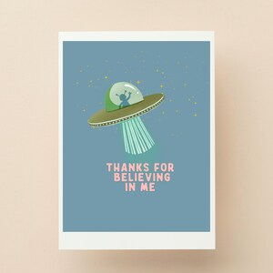 Könnte beinhalten: Eine Grußkarte mit einer Cartoon-Illustration einer grünen und goldenen UFO mit einem kleinen Alien darin, das einen Lichtstrahl aussendet. Der Text "THANKS FOR BELIEVING IN ME" ist rosa. Der Hintergrund ist blau mit gelben Sternen.
