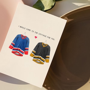 May include: Greeting card with the text "I would come to the cottage for you." Features illustrations of two hockey jerseys, one blue and one black, with a small pink heart. The card is on a cream background.