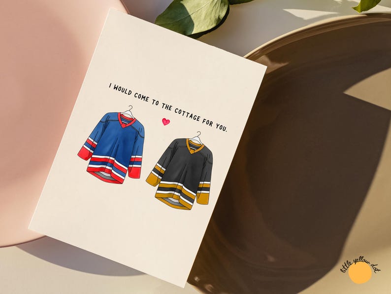 May include: Greeting card with the text "I would come to the cottage for you." Features illustrations of two hockey jerseys, one blue and one black, with a small pink heart. The card is on a cream background.
