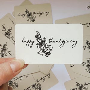 May include: Cream-colored cards with the words "happy thanksgiving" in a handwritten font. A fork and ribbon graphic is in the center. The cards are scattered, with one held in a hand.