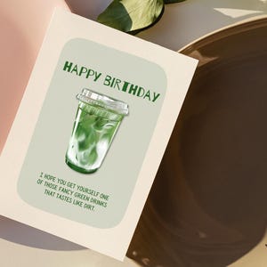 May include: A greeting card with the words "HAPPY BIRTHDAY" in green. The card features an illustration of a green drink in a clear plastic cup. The text below reads, "I HOPE YOU GET YOURSELF ONE OF THOSE FANCY GREEN DRINKS THAT TASTES LIKE DIRT."
