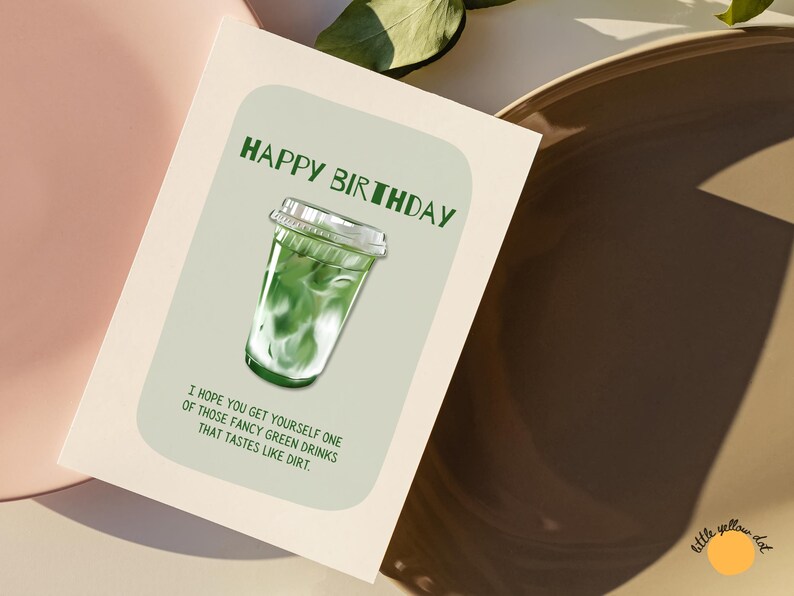May include: A greeting card with the words "HAPPY BIRTHDAY" in green. The card features an illustration of a green drink in a clear plastic cup. The text below reads, "I HOPE YOU GET YOURSELF ONE OF THOSE FANCY GREEN DRINKS THAT TASTES LIKE DIRT."
