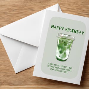 May include: A birthday greeting card with a green matcha drink illustration and the text "HAPPY BIRTHDAY". The card includes the message "I hope you get yourself one of those fancy green drinks that tastes like dirt." A white envelope is next to the card.