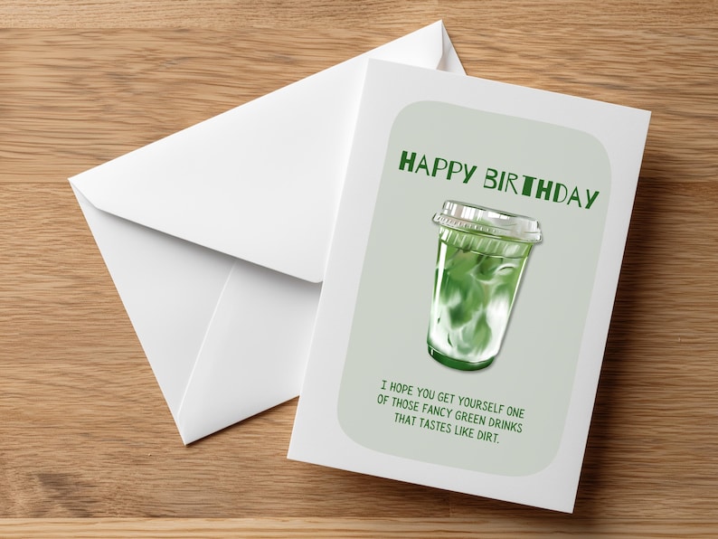 May include: A birthday greeting card with a green matcha drink illustration and the text "HAPPY BIRTHDAY". The card includes the message "I hope you get yourself one of those fancy green drinks that tastes like dirt." A white envelope is next to the card.