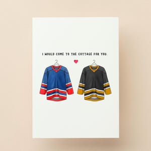 May include: Greeting card with the text "I would come to the cottage for you." Features illustrations of two hockey jerseys, one blue and red, the other black and gold, with a small pink heart.