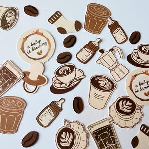 May include: A collection of die-cut paper stickers featuring coffee-themed illustrations including cups of latte art, coffee beans, baby bottles, pacifiers, socks, and circular labels with the text a baby is brewing in brown and cream tones.