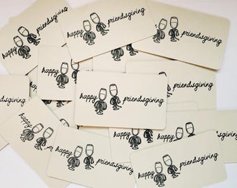 May include: A collection of off-white rectangular cards with the words "happy friendsgiving" in black script. Each card features a whimsical illustration of two wine glasses with arms and legs. The cards are scattered across a white surface.